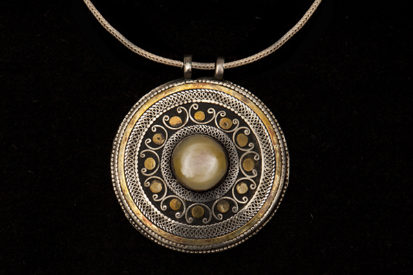 An Afghan Silvered And Golden Metal Patterned Circular Pendant With A Light Brown Domed Agate Centre