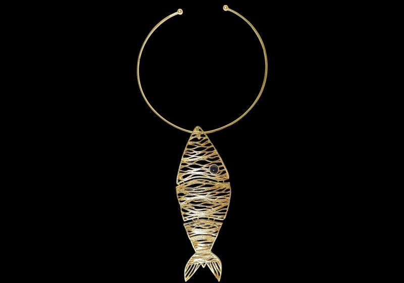 A Large Golden Metal Fish Pendant Suspended From A Golden Metal Open Circular Band Necklace.